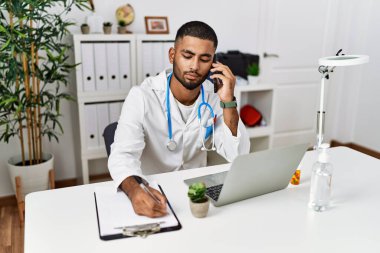 Doktor üniforması giyen genç bir ortadoğulu klinikte akıllı telefondan konuşuyor.