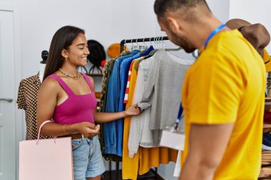 Young latin shopkeeper showing clothes to customer at clothing store.