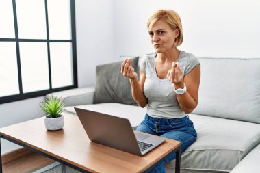 Middle age blonde woman using laptop at home doing money gesture with hands, asking for salary payment, millionaire business 