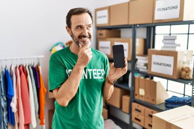 Orta yaşlı sakallı, gönüllü tişörtlü, çenesi çenesinde akıllı bir telefonla soru soran, düşünceli bir adam. Gülümseyen ve düşünceli bir yüz. şüphe kavramı. 