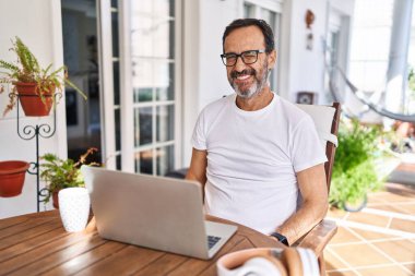 Orta yaşlı bir adam evde bilgisayar kullanıyor. Kameraya seksi bir ifadeyle bakıyor. Neşeli ve mutlu bir yüzü var.. 