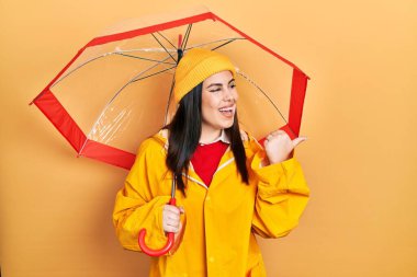 Young hispanic woman wearing yellow raincoat holding umbrella pointing thumb up to the side smiling happy with open mouth 