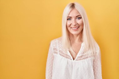 Sarı arka planda duran beyaz kadın pozitif ve mutlu görünüyor ve kendine güvenen bir gülümsemeyle dişlerini gösteriyor. 