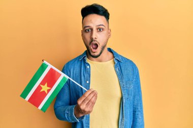 Young arab man holding suriname flag scared and amazed with open mouth for surprise, disbelief face 