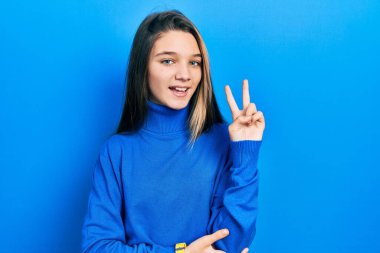 Balıkçı yaka kazak giyen ve kameraya gülümseyen genç esmer kız parmaklarıyla zafer işareti yapıyor. İki numara.. 