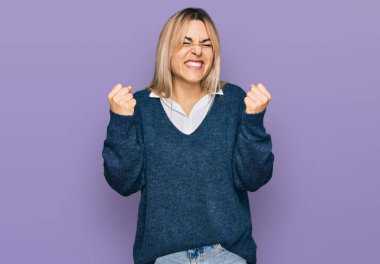 Gündelik kıyafetler giyen, mutlu ve heyecanlı genç beyaz bir kadın kolları havada, gülümseyen ve başarı için bağıran kazanan bir jest yapıyor. kutlama konsepti. 