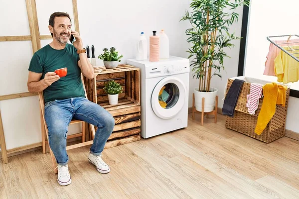 Middle Age Hispanic Man Doing Laundry Talking Smartphone Drinking Coffee — стоковое фото