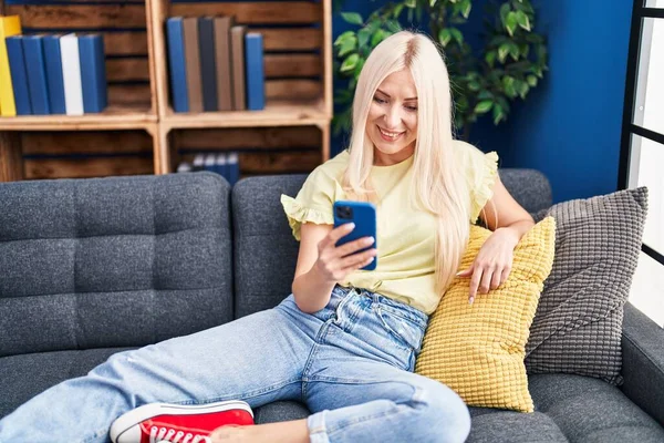 Młoda Blondynka Pomocą Smartfona Siedzi Kanapie Domu — Zdjęcie stockowe