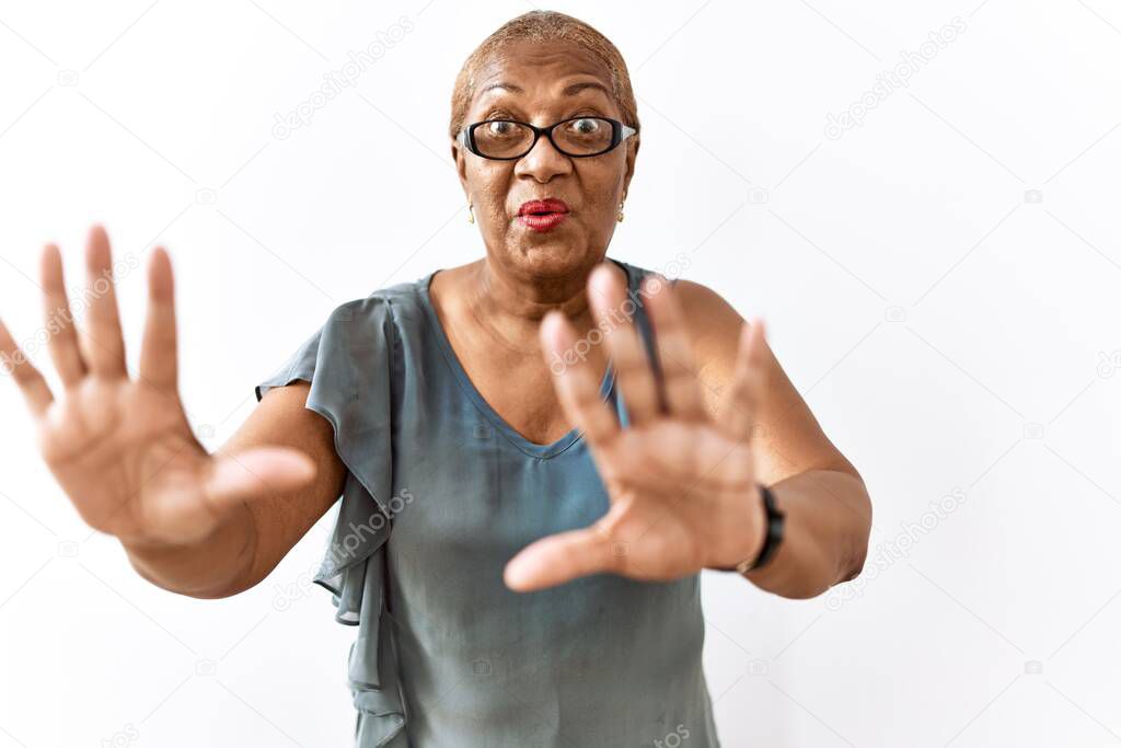 Mature hispanic woman wearing glasses standing over isolated background afraid and terrified with fear expression stop gesture with hands, shouting in shock. panic concept. 