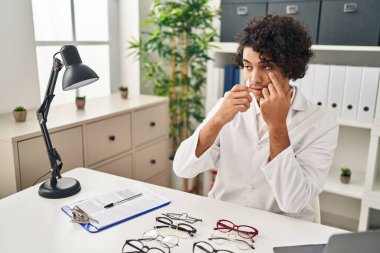 Young hispanic man optician applying eye drop at clinic