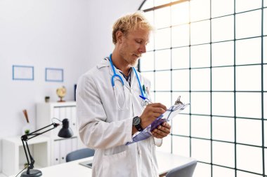 Klinikte elinde dosya tutan doktor üniforması giyen beyaz bir genç.