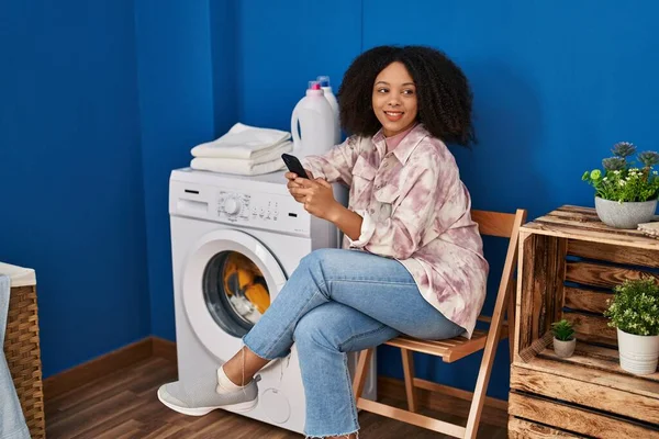Young African American Woman Using Smartphone Waiting Washing Machine Laundry — стоковое фото