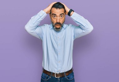 Orta yaşlı, sakallı, iş gömleği giyen deli ve elleri kafasında korkmuş, korkmuş ve ağzı açık bir şekilde şok geçirmekten korkan bir adam. 