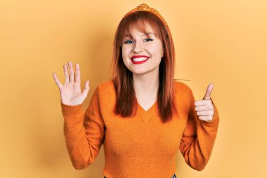 Kızıl saçlı, turuncu süveter giyen genç bir kadın altı numaralı parmaklarıyla gülümseyerek mutlu ve kendinden emin bir şekilde onu işaret ediyor.. 