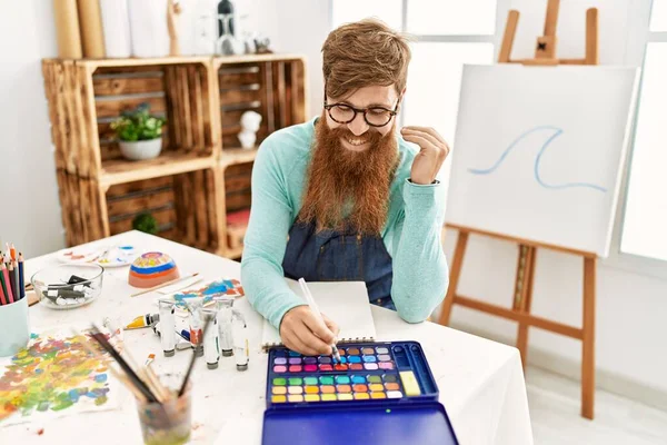 在艺术工作室 年轻的红头发男人自信地微笑着在笔记本上画画 — 图库照片