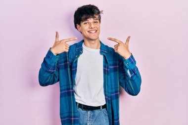 Yakışıklı hippi genç adam, günlük beyaz tişört ve klasik tişört giyiyor. Gülümsüyor, parmakları, dişleri ve ağzı ile işaret ediyor. diş sağlığı kavramı. 