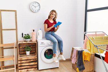 Dokunmatik ped kullanan genç beyaz bir kız evde çamaşır makinesinin başında oturmuş bekliyor..