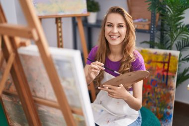 Sanat stüdyosunda kendine güvenen genç sarışın sanatçı gülümsüyor.