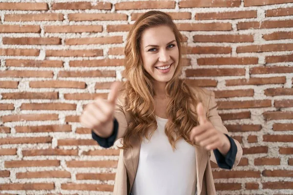 Beautiful blonde woman standing over bricks wall approving doing positive gesture with hand, thumbs up smiling and happy for success. winner gesture.
