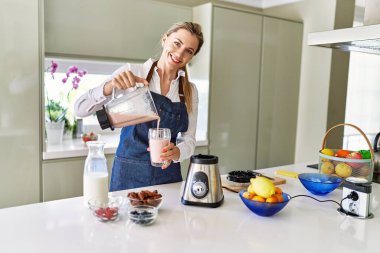 Genç sarışın kadın gülümsüyor. Kendine güvenen. Mutfaktaki cama meyve suyu döküyor.