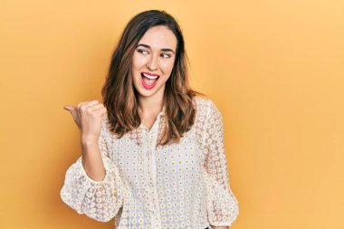 Gündelik kıyafetler giyen, gülümseyen, gülümseyen ve başparmağıyla yan tarafı gösteren İspanyol genç kız.. 