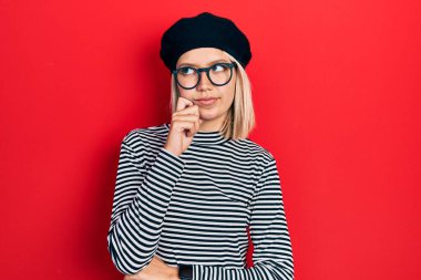Beautiful blonde woman wearing french look with beret an glasses serious face thinking about question with hand on chin, thoughtful about confusing idea 
