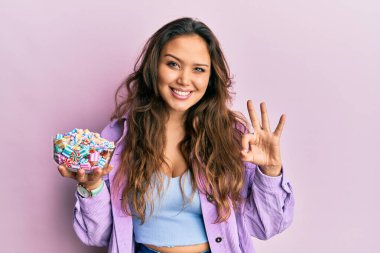 Young hispanic girl holding bowl of sugar candy doing ok sign with fingers, smiling friendly gesturing excellent symbol 