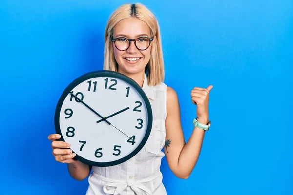 Beautiful Blonde Woman Holding Big Clock Pointing Thumb Side Smiling — 스톡 사진