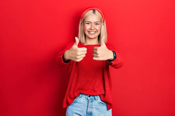 Beautiful blonde woman wearing casual red sweater approving doing positive gesture with hand, thumbs up smiling and happy for success. winner gesture.