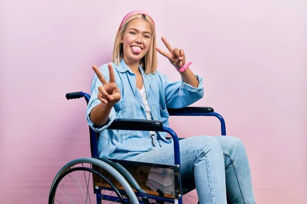 Beautiful blonde woman sitting on wheelchair smiling with tongue out showing fingers of both hands doing victory sign. number two.