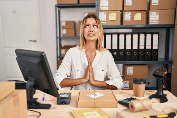 Young blonde woman working at small business ecommerce begging and praying with hands together with hope expression on face very emotional and worried. begging.
