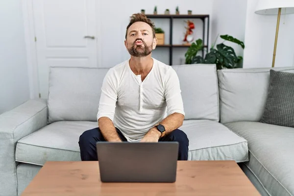 Middle age man using laptop at home looking at the camera blowing a kiss on air being lovely and sexy. love expression.