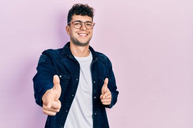 Gündelik kıyafetler giyen ve gözlüklü genç İspanyol adam mutlu ve komik yüzlü parmaklarını kameraya doğrultuyor. iyi enerji ve titreşimler. 
