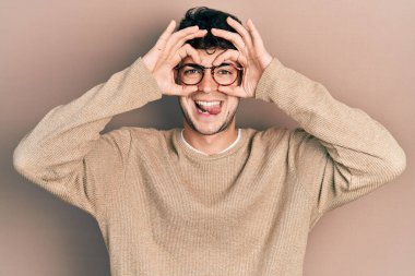 Günlük kıyafetler ve gözlük takan genç İspanyol adam dürbün gibi jest yapıyor. Dürbün gibi dilini çıkarıyor, gözler parmaklarından bakıyor. çılgın ifade. 