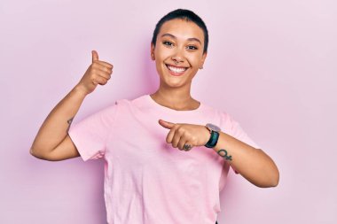 Kısa saçlı, sıradan pembe tişörtlü güzel bir İspanyol kadın. Arka tarafı işaret ediyor. El ve baş parmakları havada, kendine güvenen. 