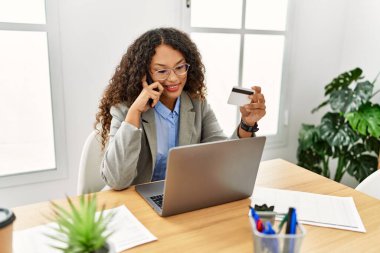 Ofiste çalışan genç Latin bir kadın elinde kredi kartıyla akıllı telefondan konuşuyor.