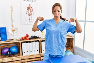 Orta yaşlı İspanyol fizyoterapist kadın ağrı kesici kliniğinde çalışıyor üzgün ve üzgün görünüyor, parmaklarıyla yön gösteriyor, mutsuz ve depresif.. 