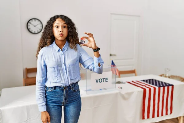 Beautiful hispanic woman standing by at political campaign by voting ballot puffing cheeks with funny face. mouth inflated with air, crazy expression.