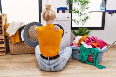 Çamaşır makinesinin yanında çamaşır yıkayan genç sarışın kadın kendine sarılıp mutlu ve pozitifmiş gibi davranıyor. Sevgi ve özgüven. 