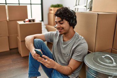 Genç İspanyol adam kulaklık ve akıllı telefon kullanıyor. Yeni evinde yerde oturuyor..