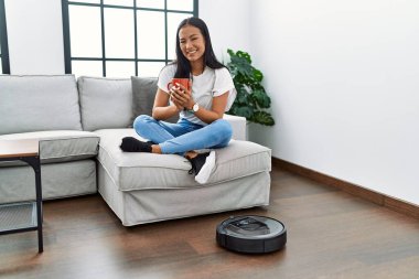 Genç Latin kadın evde temizlik robotu kullanarak kahve içiyor.