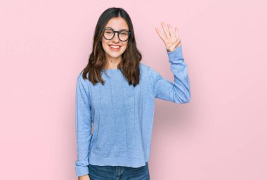 Genç ve güzel bir kadın günlük kıyafetler ve gözlük takıyor. Dört numarayı gösteriyor ve işaret ediyor. Kendinden emin ve mutlu bir şekilde gülümsüyor.. 