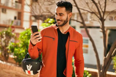 Genç İspanyol adam şehirde moto miğferi tutan akıllı telefon kullanıyor..