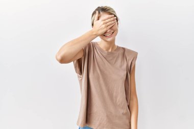 İzole edilmiş arka planda duran genç sarışın kadın. Gülümsüyor ve gülümsüyor. Gözlerini şaşkınlıkla kapatıyor. kör kavram. 