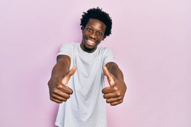 Beyaz bir tişört giyen genç Afro-Amerikan adam el hareketleriyle olumlu jest yapmayı onaylıyor, gülümsüyor ve başarı için seviniyor. kazanan jest. 