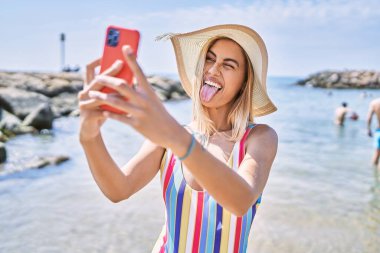 Yaz şapkası takan genç sarışın kız sahilde akıllı telefonun yanında selfie çekiyor..