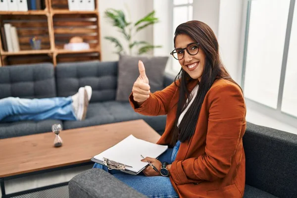 Young hispanic woman working as psychology counselor approving doing positive gesture with hand, thumbs up smiling and happy for success. winner gesture.