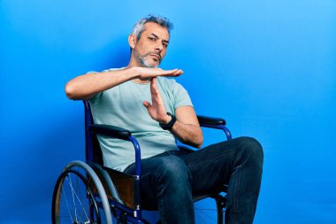 Yakışıklı, orta yaşlı, gri saçlı, tekerlekli sandalyede oturan, el hareketi yapan, hayal kırıklığına uğramış ve ciddi yüzlü bir adam. 