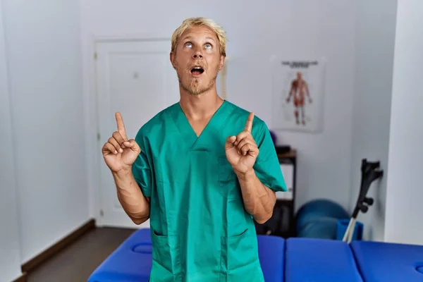 Ung Blond Man Bär Sjukgymnast Uniform Stående Kliniken Förvånad Och — Stockfoto