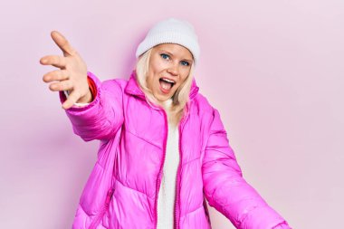 Güzel, beyaz, sarı saçlı, yün şapkalı, kışlık ceketli bir kadın kameraya bakıyor. Sarılmak için kollarını açmış gülümsüyor. Mutluluğu kucaklayan neşeli bir ifade.. 